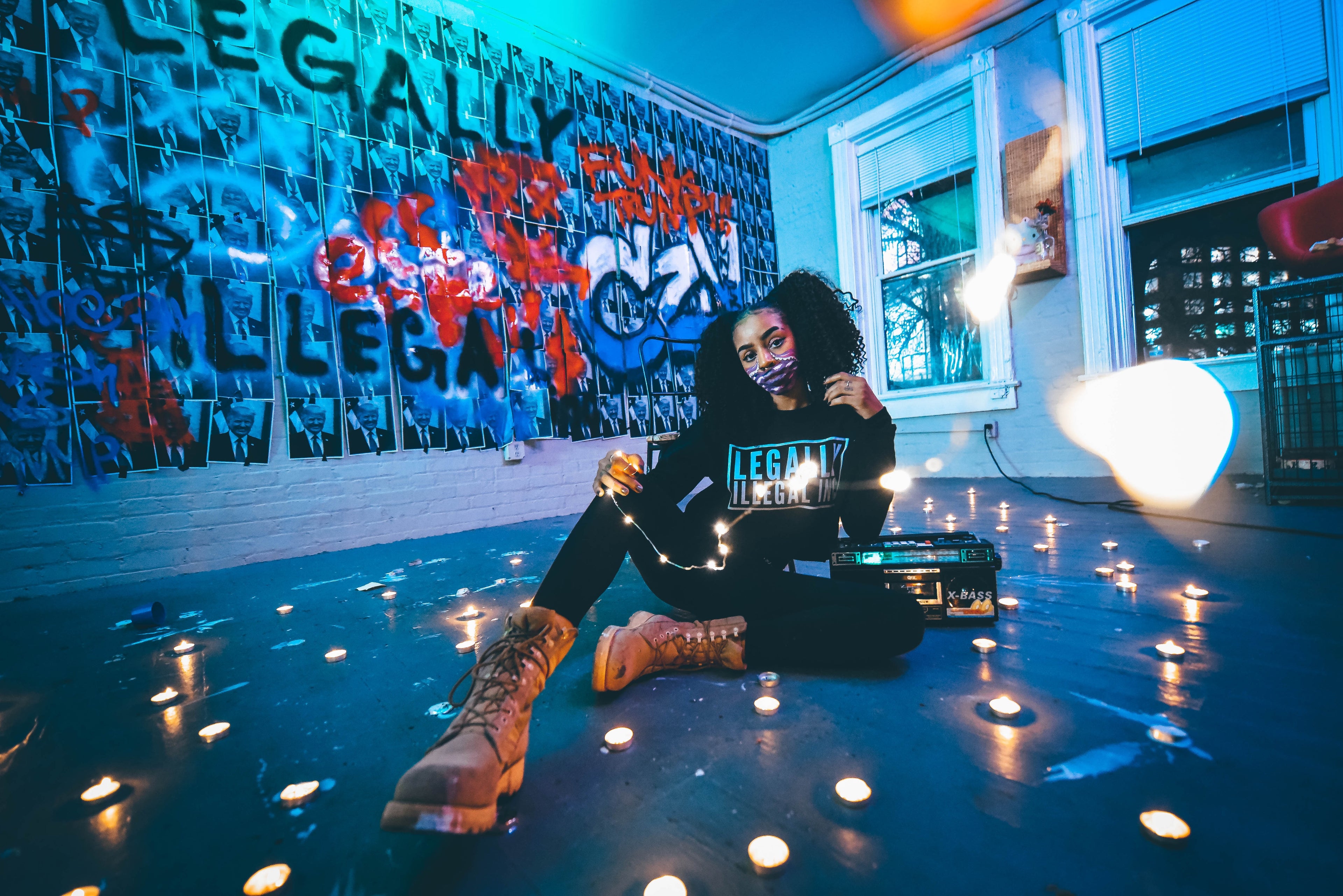 Person sitting on a floor with candles, wearing a 'Legally Illegal Black' sweatshirt in a room with graffiti on the wall.